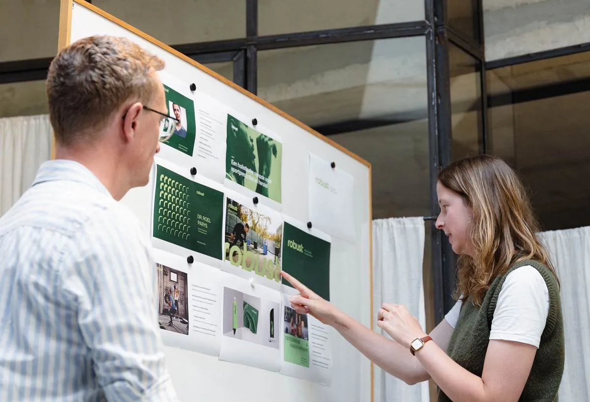 Twee marketeers bespreken een marketingstrategie en analyseren visuele plannen op een presentatiebord.