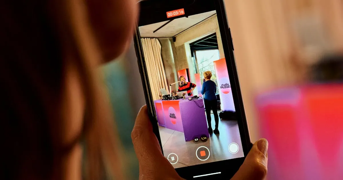 Persoon filmt met een smartphone een presentator achter een kleurrijke desk in een binnenruimte.