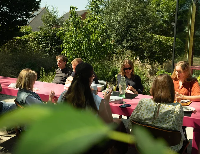 Team zit samen aan een lange tafel buiten op kantoor en luncht in een groene tuinomgeving.